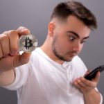 Homem jovem olhando para celular e segurando um Bitcoin criptomoedas FIRE