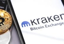 Celular com aplicativo da corretora Kraken ao lado de moeda do Bitcoin