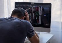 Homem jovem desesperado com as mãos na cabeça em frente ao computador com gráficos do bitcoin