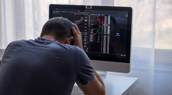 Homem jovem desesperado com as mãos na cabeça em frente ao computador com gráficos do bitcoin
