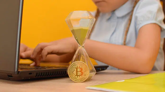 Criança próxima a um notebook, ampulheta e moeda de Bitcoin