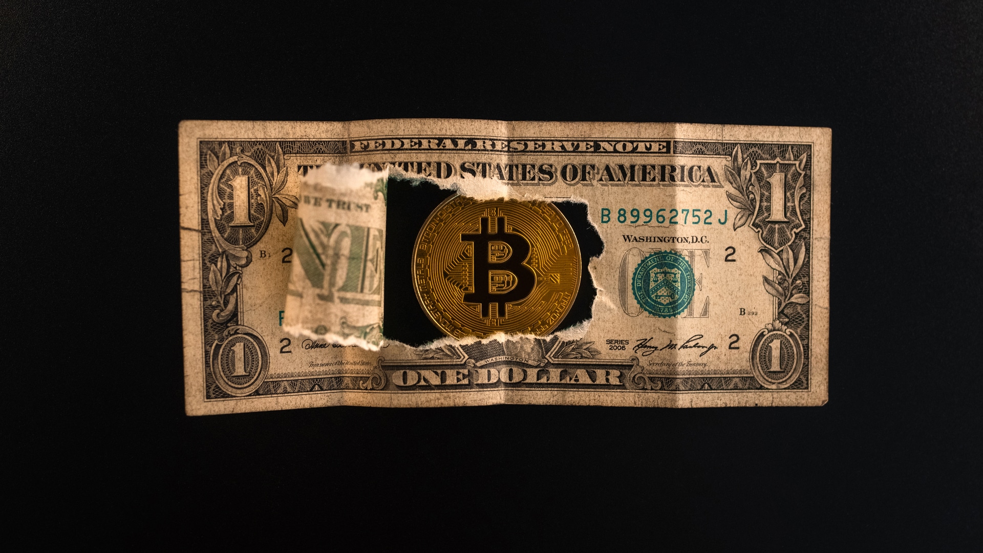 Uma nota de um dólar rasgada através da qual as moedas Bitcoin são visíveis