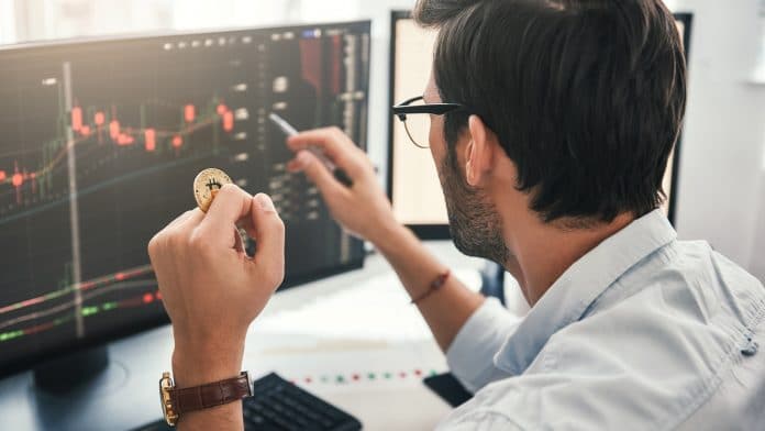 Jovem trader apontando para os dados na tela do computador com caneta e segurando Bitcoin