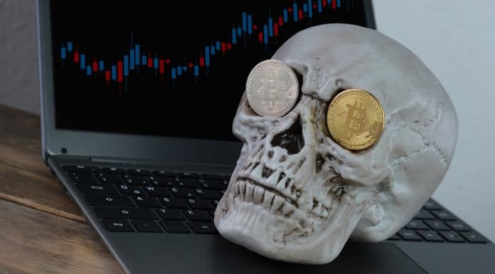Esqueleto com Bitcoin nos olhos próximo de computador com gráficos