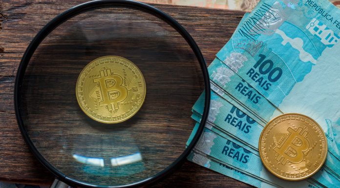Lupa focando em moeda física de Bitcoin ao lado de notas de 100 reais.