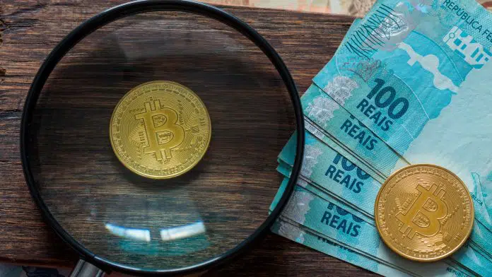 Lupa focando em moeda física de Bitcoin ao lado de notas de 100 reais.
