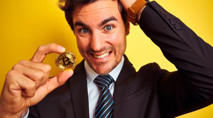 Homem segurando moeda de Bitcoin com cara de confuso.