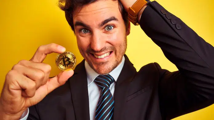 Homem segurando moeda de Bitcoin com cara de confuso.