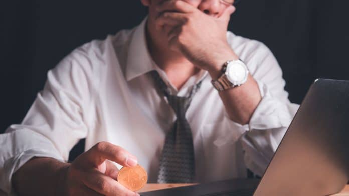 Homem triste enquanto segura moeda de Bitcoin enquanto olha para computador.
