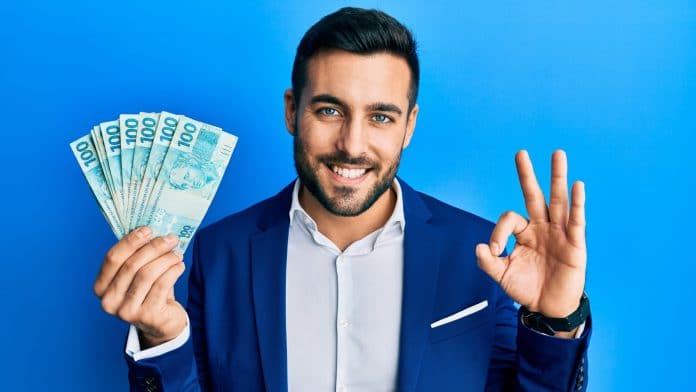 Empresário vestindo terno segurando notas reais brasileiras fazendo sinal ok