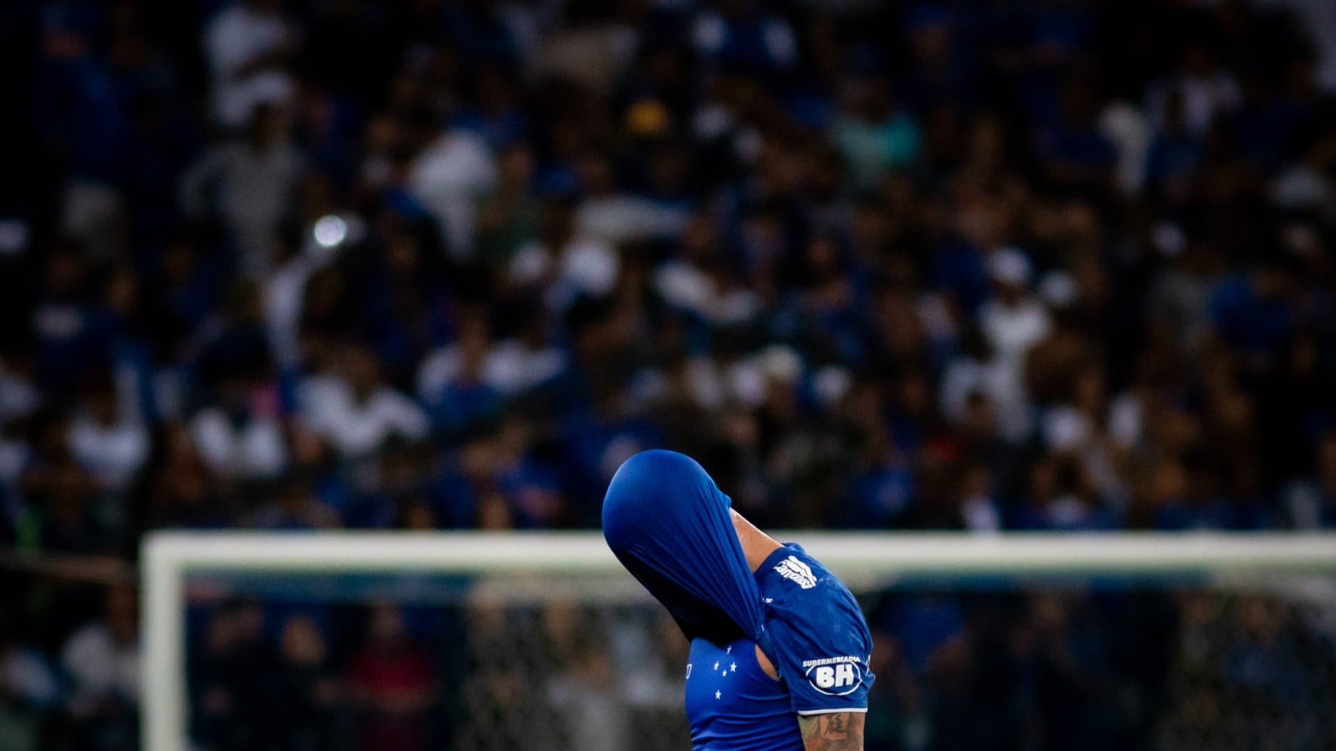 Jogador do Cruzeiro tampando cabeça com camisa durante partida de futebol em estádio