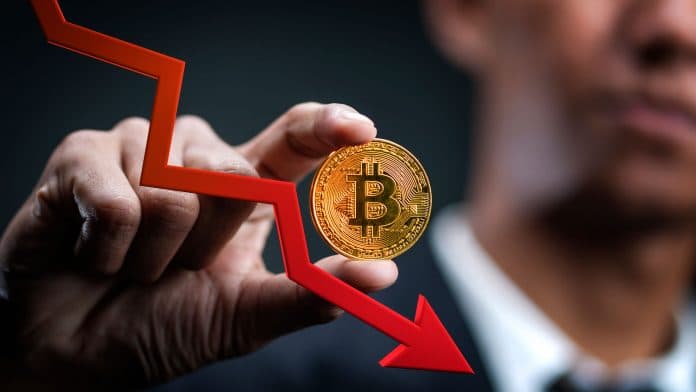 Homem segurando moeda de Bitcoin (BTC) e seta apontando queda do mercado.
