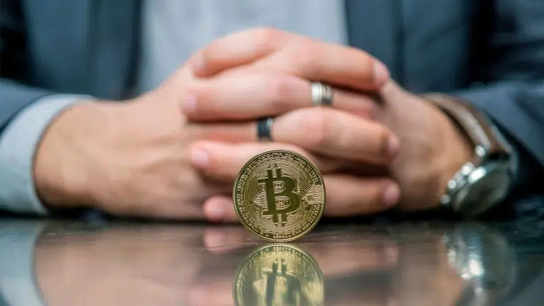Moeda de Bitcoin parada sobre a mesa, em frente a mãos de empresário.