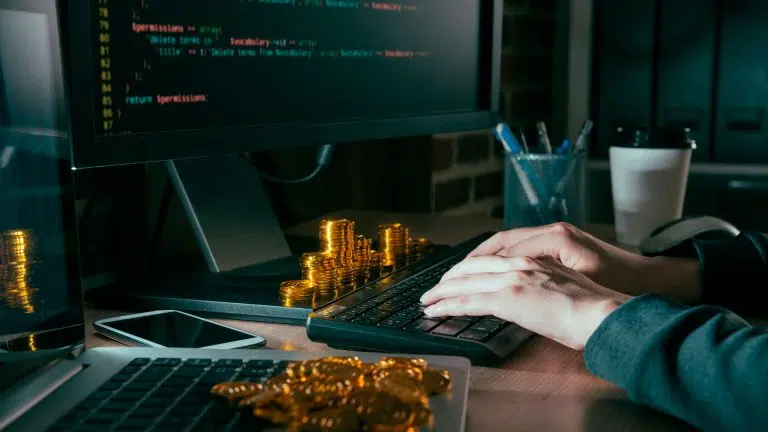 Hacker escrevendo código ao lado de pilha de criptomoedas.