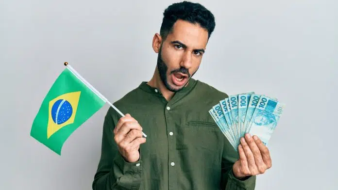 Jovem segurando a bandeira do brasil e notas de reais com rosto em choque após calote com criptomoedas