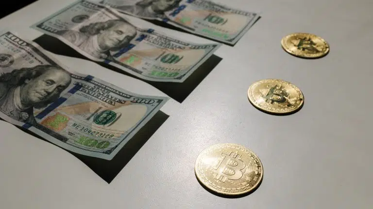 Moedas físicas de Bitcoin em frente a notas de 100 dólares.