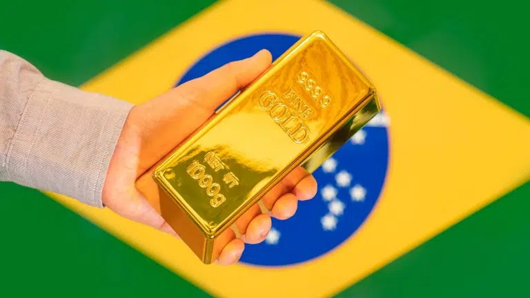 Mão segurando barra de ouro com bandeira do Brasil ao fundo