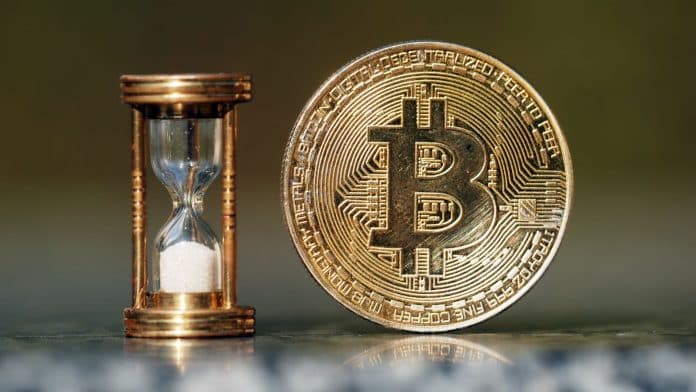 Moeda física de Bitcoin ao lado de ampulheta.