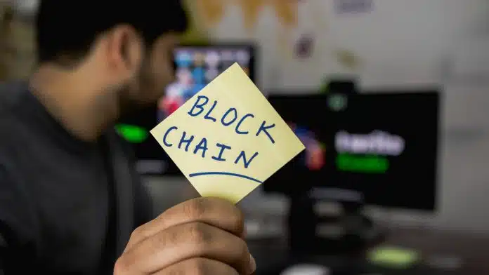 Homem segurando papel escrito blockchain enquanto observa tela de monitor