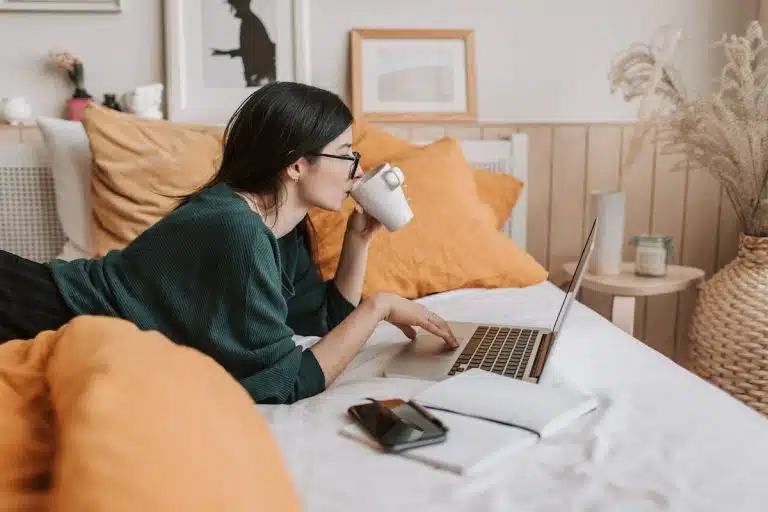 Mulher estudando em computador enquanto toma café e deitada na cama, concurso público
