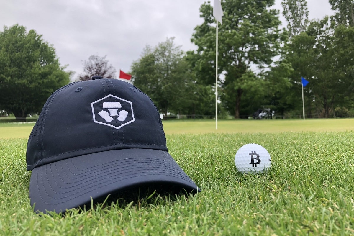 Boné com a marca da corretora Crypto ao lado de bola de golfe com símbolo do bitcoin