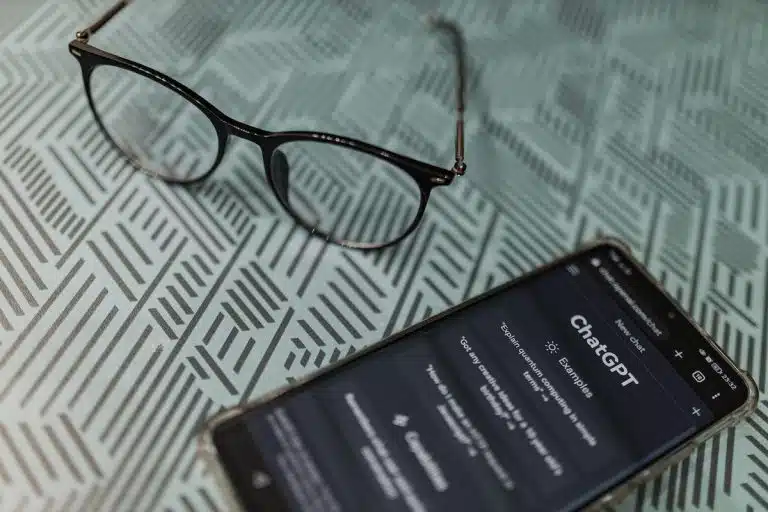 Óculos em mesa de ferro ao lado de celular com imagem do ChatGPT