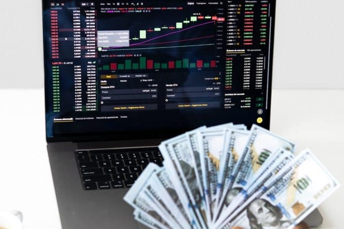 Homem segurando notas de dinheiro em frente a notebook com tela gráfica de preços de criptomoedas