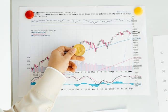 Mão segurando bitcoin em frente a gráficos de preços