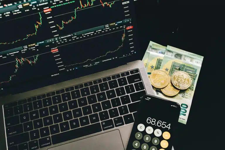 Moedas de bitcoin sobre notas de Euro, ao lado de computador em tela de negociações