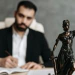 Homem ofuscado ao fundo, observando estátua da justiça sobre a mesa