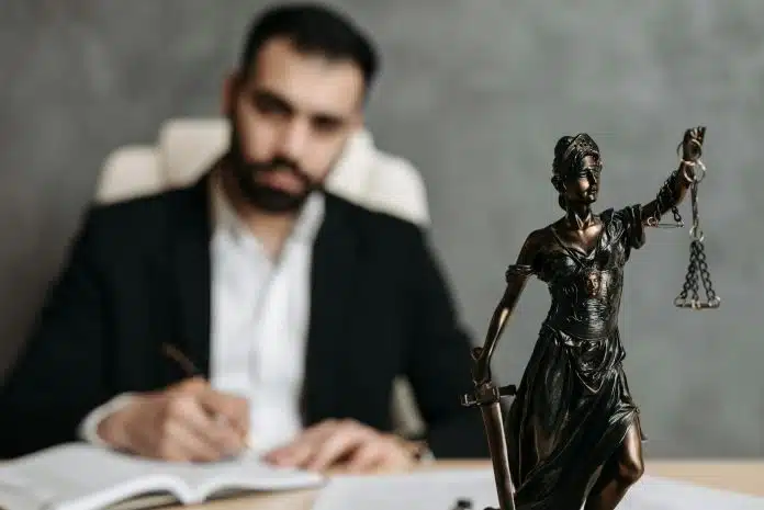 Homem ofuscado ao fundo, observando estátua da justiça sobre a mesa