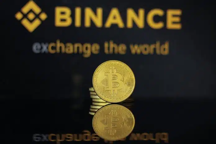 Logo da Binance ao fundo, com bitcoin simbólico a frente