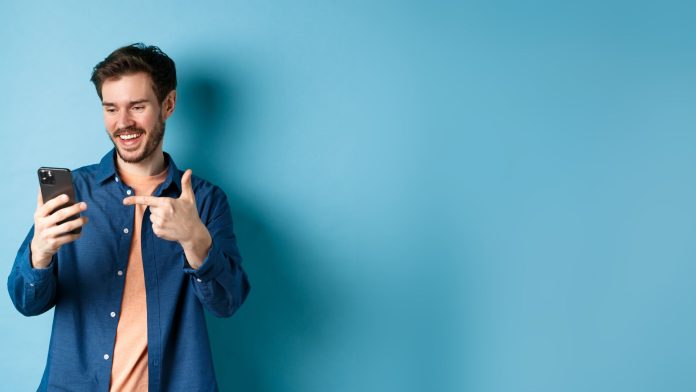 Homem com fundo azul sorrindo ao apontar para celular aplicativo Telegram