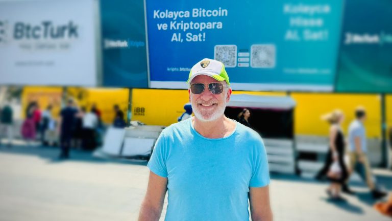 Peter Schiff sorrindo com placa de bitcoin aos fundos