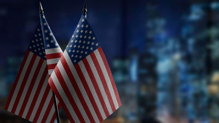 Bandeira dos Estados Unidos em destaque com prédios ao fundo FBI revela perdas bilionárias com fraudes com criptomoedas e alerta investidores sobre táticas de inteligência artificial