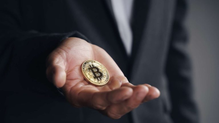 Homem de terno segurando bitcoin em destaque Governo proíbe uso de criptomoedas no eFX; o que muda?