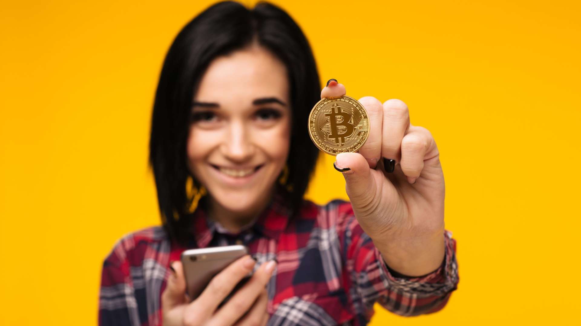 Mulher segurando bitcoin e celular sorrindo