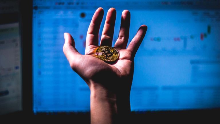 Mão segurando bitcoin em frente a tela de computador